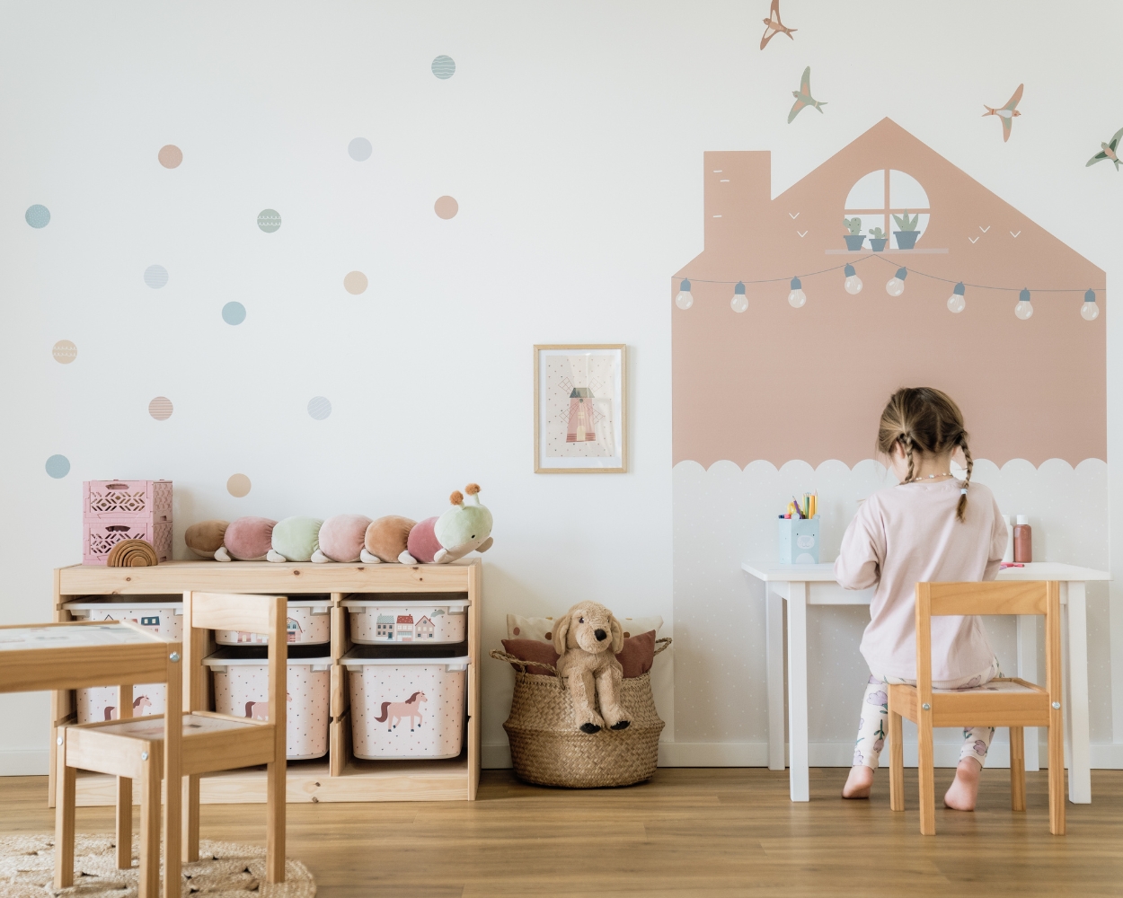 Ein Kinderzimmer für Mädchen mit rosa Wandtattoos, einem Ikea Trofast Regal mit Klebefolien mit Pferden, ein Tisch und ein Mädchen, welches an ihrem Schreibtisch stzt